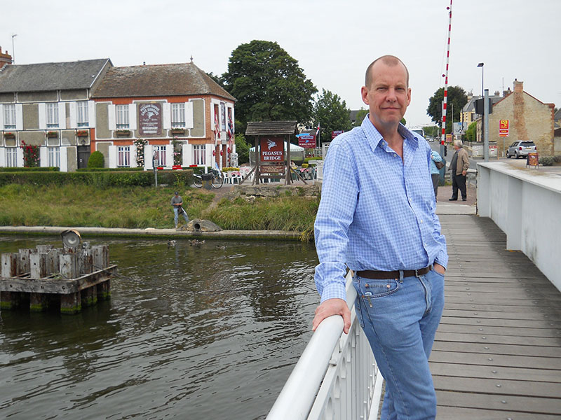 Man standing on a bridge with a river and buildings in the background.